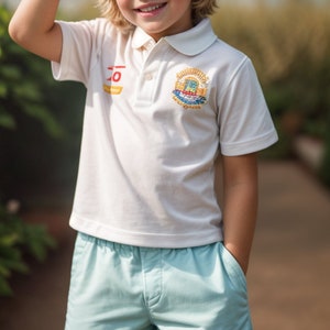 Puede incluir: Un ni&ntilde;o peque&ntilde;o con una camiseta polo blanca con un logotipo bordado y pantalones cortos de color azul claro. El logotipo presenta un dise&ntilde;o colorido con un edificio y el texto "Junior Sailing Club".