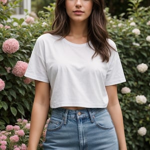 Puede incluir: Una mujer con una camiseta blanca recortada y unos vaqueros de cintura alta de color azul claro. Está de pie frente a un arbusto verde con flores rosas.