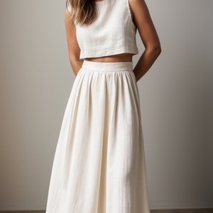 May include: A cream-coloured two-piece outfit comprising a sleeveless crop top and a high-waisted, flowing maxi skirt. The fabric appears to be a textured linen or similar material. The outfit is simple and elegant, suitable for warm weather.