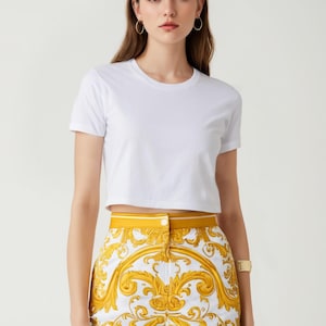 Puede incluir: Una camiseta blanca recortada combinada con pantalones cortos estampados en amarillo y blanco. Los pantalones cortos tienen un estampado barroco dorado.