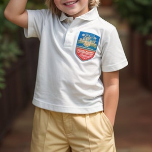 Puede incluir: Una persona joven lleva una camiseta polo blanca con un parche bordado azul y rojo que dice "Franklinville Academy" y pantalones cortos color caqui.