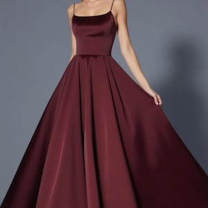 Puede incluir: Un vestido de fiesta burdeos largo hasta el suelo, con escote cuadrado y tirantes finos. El vestido presenta un corpiño ajustado y una falda amplia y fluida. La tela tiene un brillo sutil, y el diseño general es elegante y clásico.