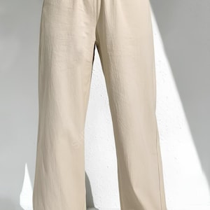 May include: Beige wide-leg trousers with an elasticated waistband. The trousers are made of a lightweight fabric and have a relaxed fit. The model is wearing white trainers. The trousers are a neutral colour, suitable for various outfits.