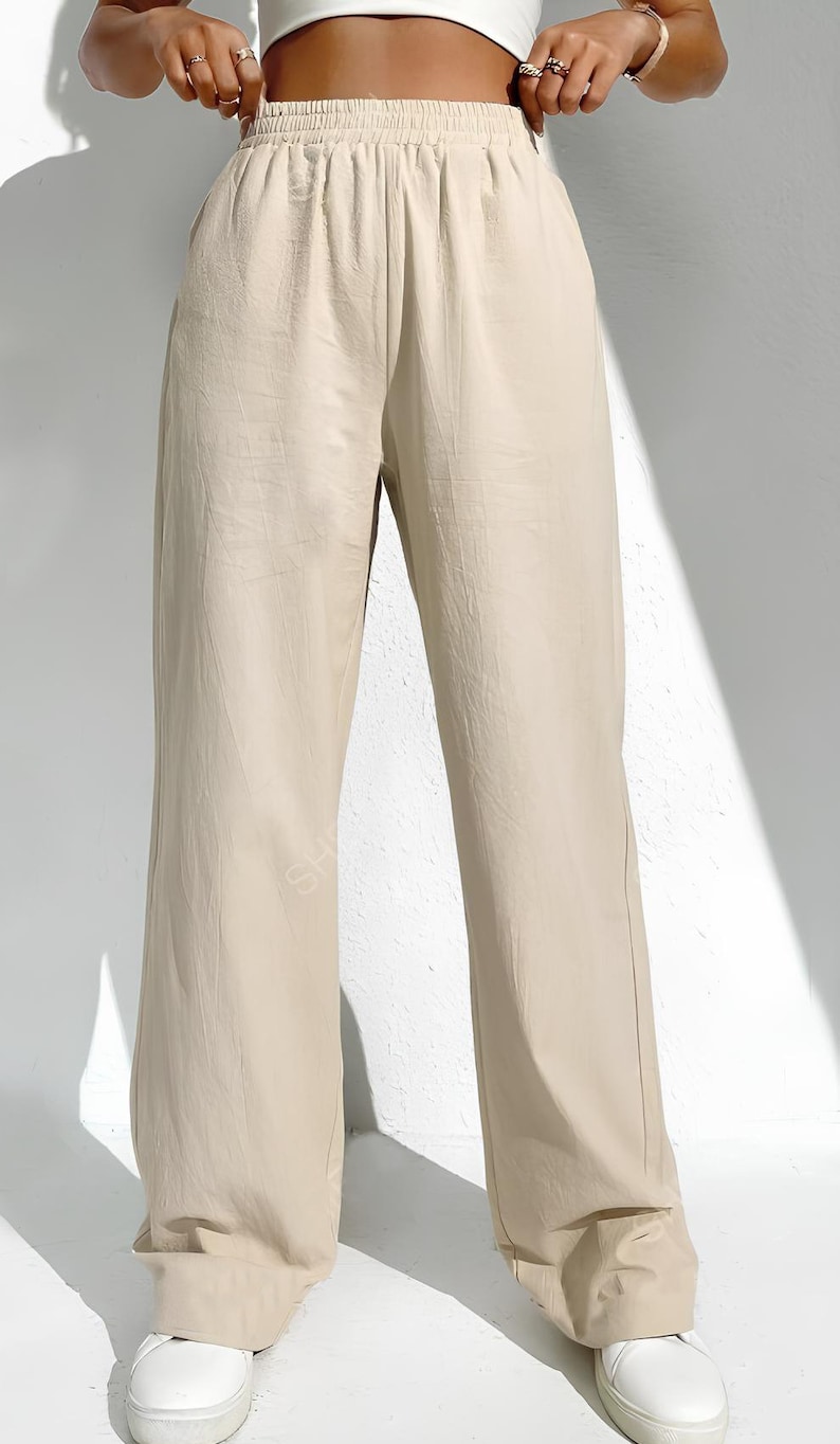 May include: Beige wide-leg trousers with an elasticated waistband. The trousers are made of a lightweight fabric and have a relaxed fit. The model is wearing white trainers. The trousers are a neutral colour, suitable for various outfits.