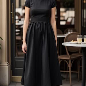 Puede incluir: Una mujer con un vestido midi negro con escote barco y mangas cortas. El vestido tiene un corpiño ajustado y una falda completa. Está de pie frente a un café.