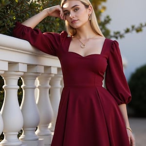 May include: A woman wearing a maroon dress with a sweetheart neckline and puff sleeves. The dress has a fitted bodice and a flowing skirt. She is standing in front of a white railing.