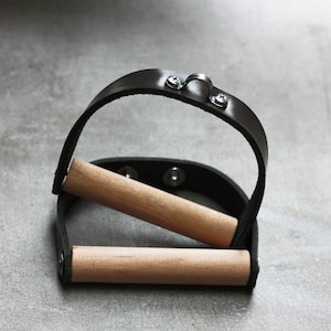 Pode incluir: Uma alça de exercício de couro preto com duas barras de madeira. A alça tem um laço de metal na parte superior.