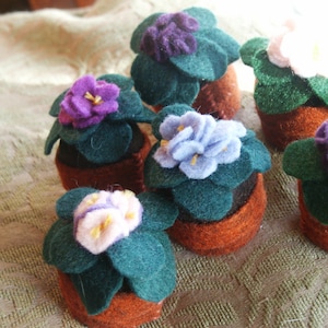 May include: A collection of felt African violet plants in small pots. The plants have green leaves and purple, blue, and pink flowers.