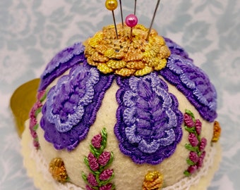 May include: A white pincushion with purple and pink embroidered flowers and a yellow flower on top. The pincushion has a white lace trim and is decorated with five colorful pins.