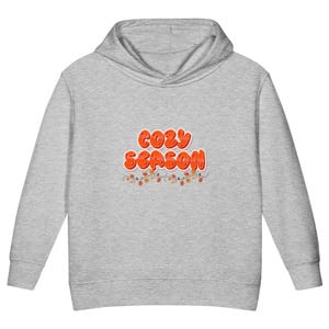 Może przedstawiać: Bluza z kapturem w kolorze szarego melanżu z napisem "COZY SEASON" wykonanym pogrubioną, pomarańczową czcionką w stylu bąbelkowym. Poniżej tekstu znajduje się ozdobna grafika przedstawiająca cienkie, wijące się gałązki z małymi jesiennymi liśćmi.