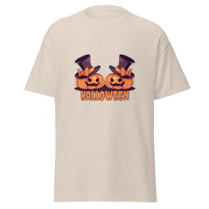 May include: A beige short-sleeve t-shirt featuring a graphic of two orange jack-o'-lanterns wearing black top hats with orange bows. Below the pumpkins, the word HALLOWEEN is printed in bold orange capital letters with a black outline.