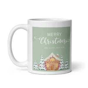 May include: A white ceramic mug featuring a sage green rectangular graphic. The design displays a gingerbread house surrounded by snow-covered pine trees and the white text Merry Christmas and Happy New Year in a script and sans-serif font.