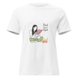 May include: A white short-sleeved t-shirt featuring a line art graphic of a person sitting cross-legged while reading a red book, accompanied by a bowl of snacks and a bubble tea. The text reads Read. Snack. Repeat. in a black cursive script font.