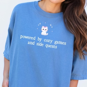 May include: A blue t-shirt with the text "powered by cozy games and side quests" and a cartoon cat graphic. The shirt is a solid blue color and has short sleeves. The words "BLUE JEAN" are in the top left corner.