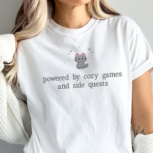 May include: White t-shirt with the text "powered by cozy games and side quests" and a gray cat graphic wearing a pink bow. The shirt is part of a casual wear set.