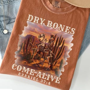 May include: A rust-colored t-shirt featuring a graphic of two skeletons riding horses through a desert landscape with cacti. The text reads DRY BONES COME-ALIVE EZEKIEL 37:4. The shirt is paired with light blue denim jeans, a tan wide-brim hat, and a necklace.