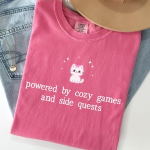 May include: A pink t-shirt with the text "powered by cozy games and side quests" and a cartoon cat graphic. The shirt is paired with blue jeans and a tan hat. The text "CRUNCH BERRY" is in the top left corner.