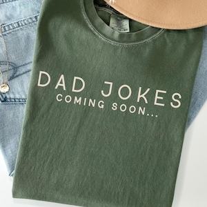 May include: A moss green crew neck t-shirt with the text DAD JOKES COMING SOON printed in a light cream color across the chest. The shirt features a visible brand label at the inner neckline and is displayed alongside light blue denim fabric.