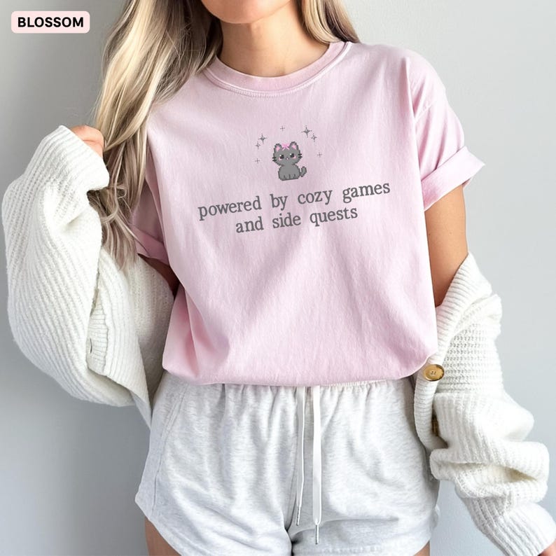 May include: A light pink t-shirt with the text "powered by cozy games and side quests" and a cartoon cat graphic. The shirt is paired with gray shorts and a white cardigan.