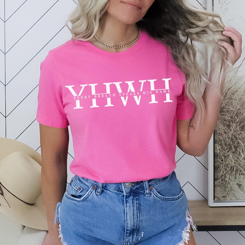K&ouml;nnte beinhalten: Ein leuchtend pinkfarbenes T-Shirt mit dem wei&szlig;en Text "VIIIIII EVERY BREATH SPEAKS HIS NAME IIIVVII". Das kurz&auml;rmelige Oberteil wird mit Jeansshorts getragen. Eine goldene Halskette ist ebenfalls zu sehen.