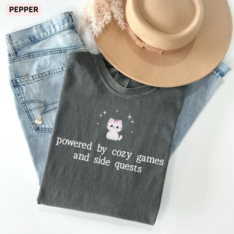 May include: A flat lay featuring a gray t-shirt with a cat graphic and the text "powered by cozy games and side quests." The shirt is paired with light blue jeans, a tan hat, and a gold necklace.