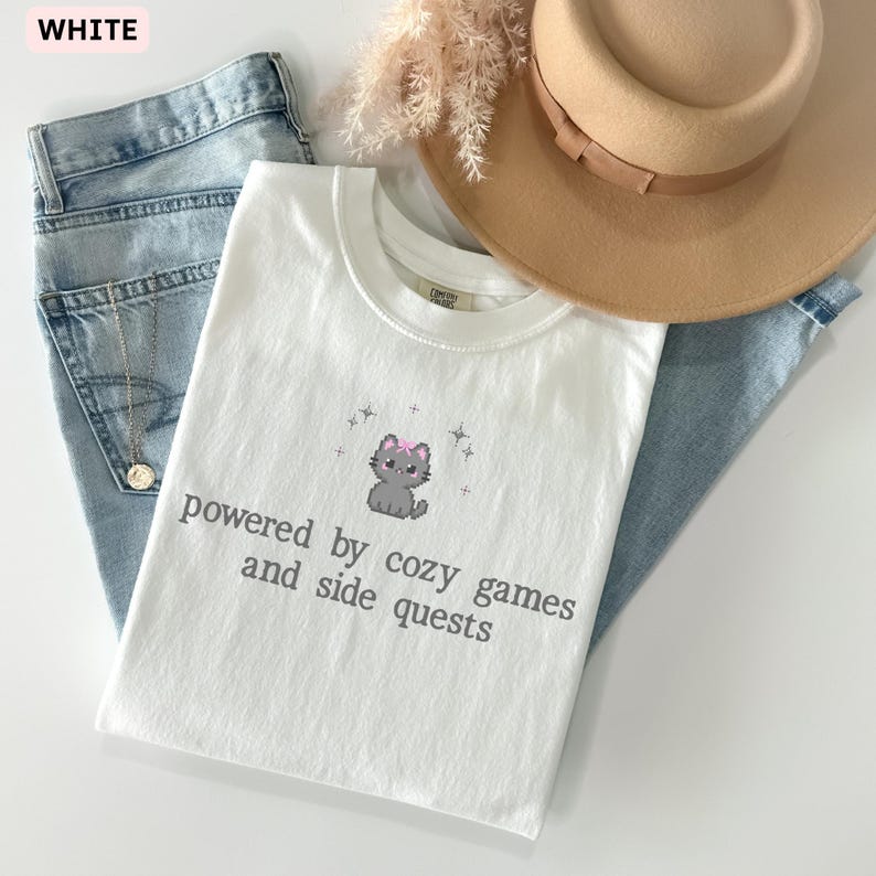 May include: White t-shirt with a graphic of a gray cat wearing a pink bow and the text "powered by cozy games and side quests." The shirt is paired with light blue jeans and a tan hat.