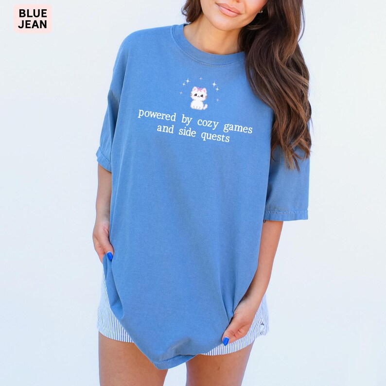 May include: A blue t-shirt with the text "powered by cozy games and side quests" and a cartoon cat graphic. The shirt is a solid blue color and has short sleeves. The words "BLUE JEAN" are in the top left corner.