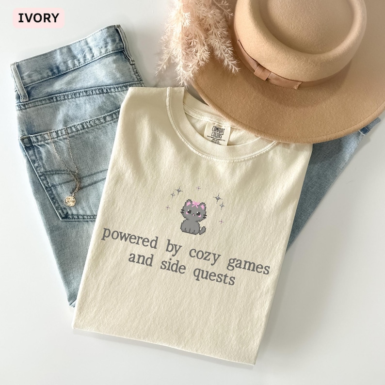 May include: Ivory-colored t-shirt with a graphic of a gray cat wearing a pink bow, with the text "powered by cozy games and side quests." The shirt is paired with light blue jeans, a tan hat, and a necklace.