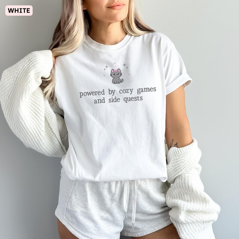 May include: White t-shirt with the text "powered by cozy games and side quests" and a gray cat graphic wearing a pink bow. The shirt is part of a casual wear set.