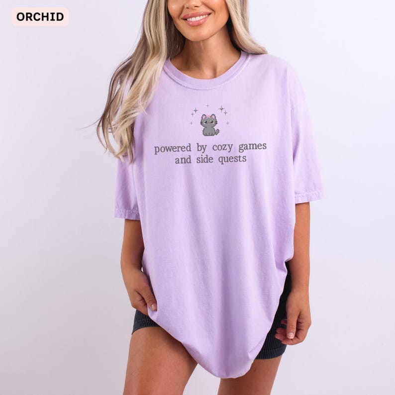 May include: A light purple t-shirt with the text "powered by cozy games and side quests" and a small gray cat graphic. The word "ORCHID" is in the upper left corner. The shirt is a comfortable, casual style.