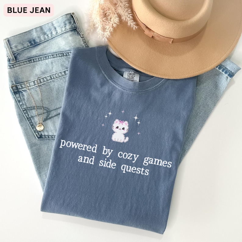May include: A blue Comfort Colors t-shirt with a white cat graphic and the text "powered by cozy games and side quests." The shirt is paired with light blue jeans and a tan hat.