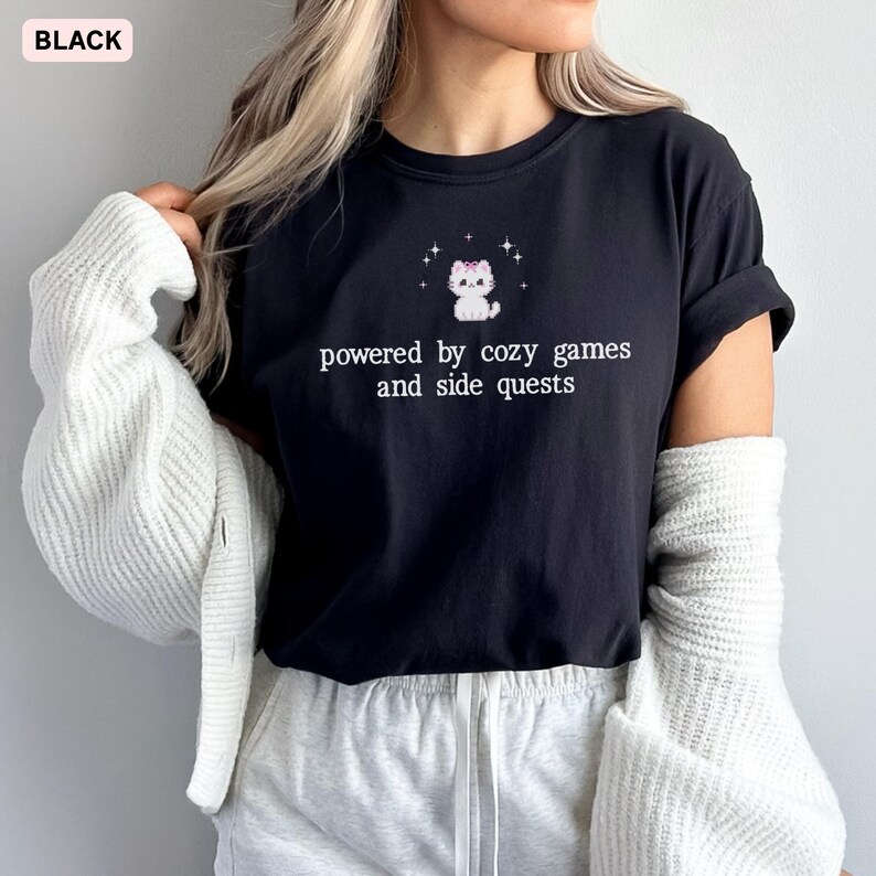 May include: Black t-shirt with the text "powered by cozy games and side quests" and a cartoon cat graphic. The cat has a pink bow and is surrounded by stars. The shirt is a casual style.