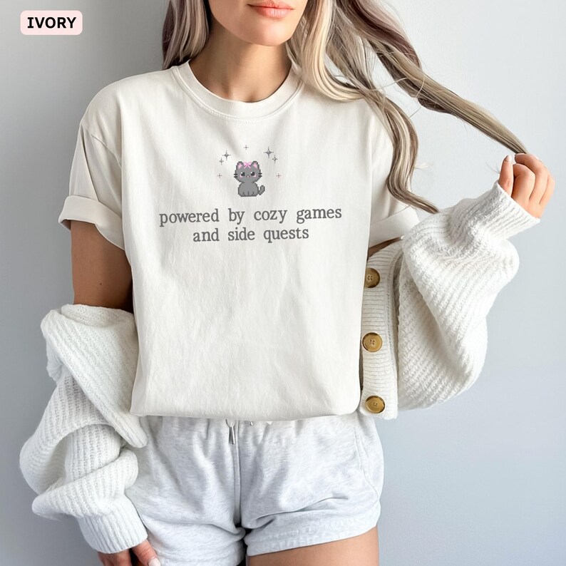 May include: Ivory-colored t-shirt with the text "powered by cozy games and side quests" and a cartoon cat graphic. The shirt is paired with gray shorts and a white cardigan.