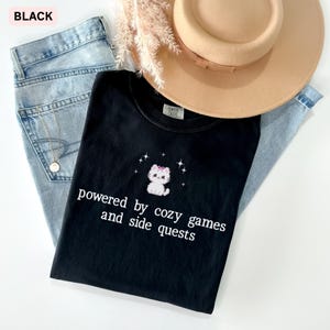 May include: Black t-shirt with a white graphic of a cat wearing a bow and the text "powered by cozy games and side quests." The shirt is paired with light blue jeans and a tan hat.