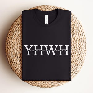 K&ouml;nnte beinhalten: Schwarzes T-Shirt mit dem wei&szlig;en Text "YHWH EVERY BREATH SPEAKS HIS NAME". Das Shirt ist gefaltet und auf einer gewebten, kreisf&ouml;rmigen Oberfl&auml;che platziert. Das Shirt hat ein Etikett mit der Aufschrift "Canvas".