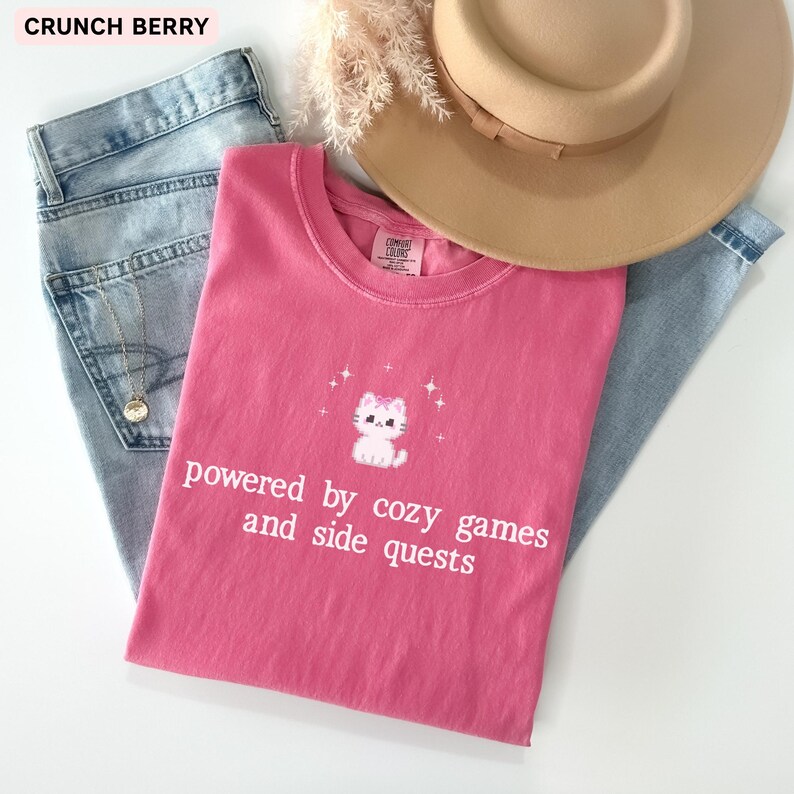 May include: A pink t-shirt with the text "powered by cozy games and side quests" and a cartoon cat graphic. The shirt is paired with blue jeans and a tan hat. The text "CRUNCH BERRY" is in the top left corner.