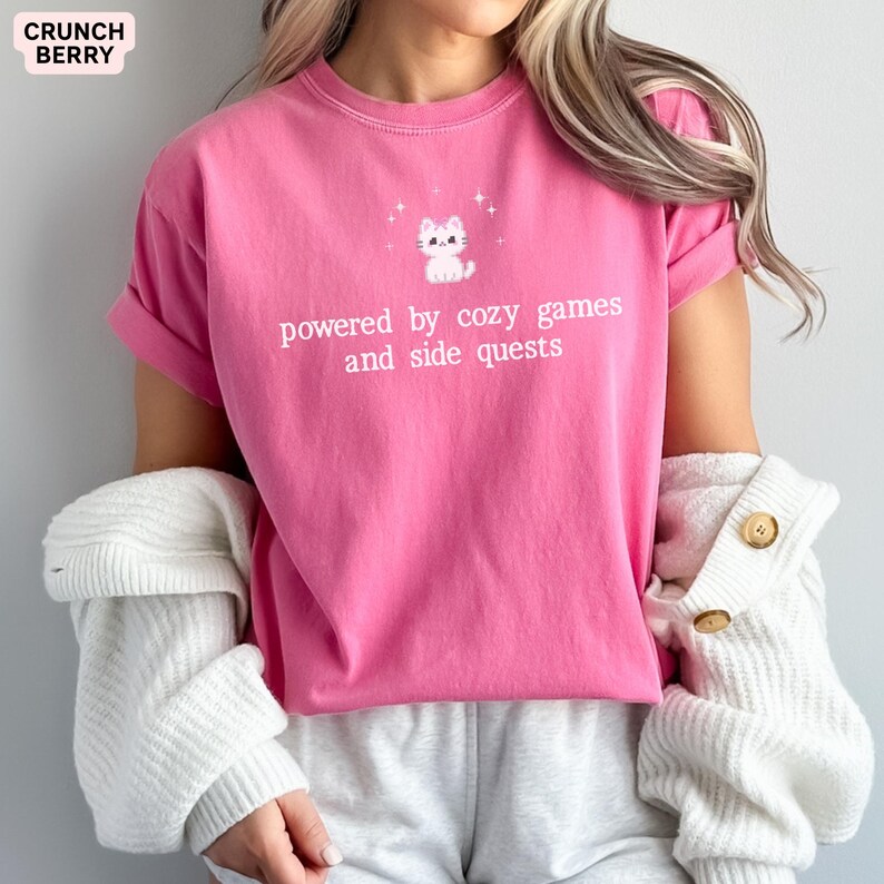 May include: A bright pink t-shirt with rolled-up sleeves. The shirt features a white graphic of a cat and the text "powered by cozy games and side quests." The brand name "Crunch Berry" is in the upper left corner.