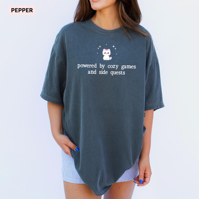 May include: Dark gray t-shirt with the text "powered by cozy games and side quests" and a small cartoon cat graphic. The shirt is a relaxed fit and has short sleeves.