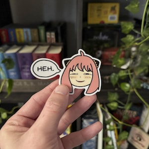 May include: A sticker featuring a cartoon character with pink hair and black cat ears. The character is smiling and has a speech bubble that says "HEH."