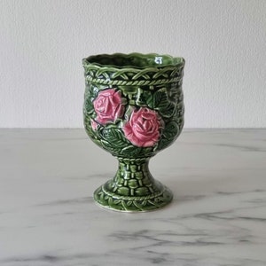 Puede incluir: Un macetero de cerámica verde con base de pedestal. El macetero está decorado con rosas rosas y hojas verdes.