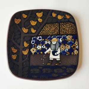 May include: A square ceramic tile with rounded corners featuring a dark brown tree with yellow birds, a figure in a white tunic scattering seeds, a blue floral background, and a dark blue river with fish at the bottom, marked with the year 1976.