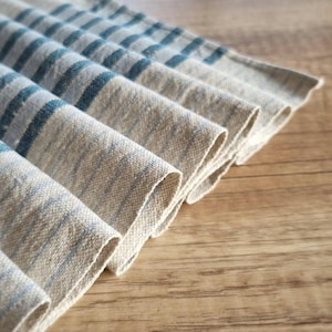 Könnte beinhalten: Gefalteter Leinenstoff mit blauen Streifen. Der Stoff hat ein natürliches, strukturiertes Aussehen und ist diagonal angeordnet. Die blauen Streifen sind gleichmäßig über den Stoff verteilt.