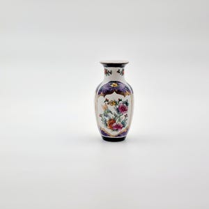 Puede incluir: Pequeño jarrón decorativo de cerámica con base blanca y diseño floral en rosa, naranja y morado. El jarrón tiene un ribete morado oscuro y dorado en la parte superior e inferior, y un borde blanco. Aproximadamente 15 cm de altura.