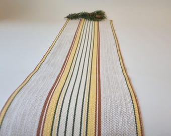 May include: A rectangular table runner with vertical stripes in cream, yellow, brown, and green. The runner is made of woven fabric and has a decorative sprig of greenery at the top.