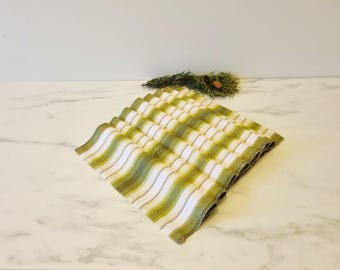 May include: A folded, square, striped cloth napkin with a wavy pattern. The stripes alternate between white, light green, and a darker olive green. The napkin is on a white marble surface, with a sprig of greenery in the background.