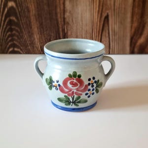Pode incluir: Um pequeno pote de cerâmica azul e branco com um design floral. O pote tem duas alças e apresenta uma rosa rosa com folhas verdes e flores azuis e amarelas.