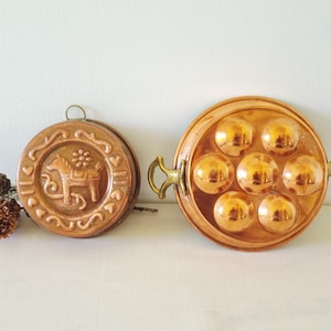 May include: Two copper kitchen molds. One is round with a horse design, the other is a round pan with seven spherical indentations. Both have a shiny, reflective surface and are likely vintage kitchenware.