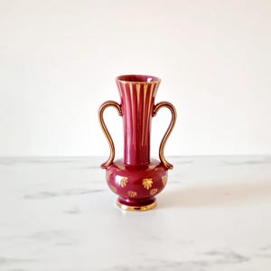 May include: A small, burgundy ceramic vase with gold accents and two handles. The vase has a ribbed design and is decorated with gold leaf patterns.