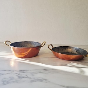 Peut inclure: Deux récipients de cuisson en cuivre avec des intérieurs métalliques et des poignées en laiton de chaque côté. Le récipient de gauche est plus profond et cylindrique, tandis que celui de droite est plus plat et de forme ovale.
