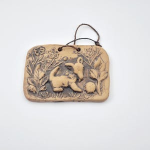 May include: A ceramic wall hanging with a brown glaze features a relief sculpture of two playful animals in a garden setting. The animals are stylized and appear to be a dog and a cat. The background is filled with leaves and flowers.